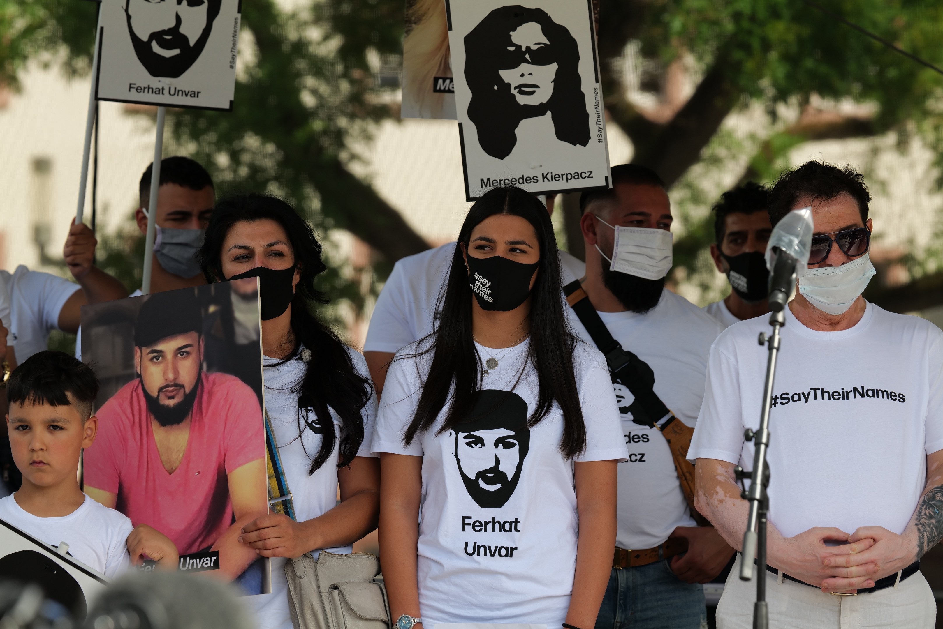 Eine Gruppe von Menschen mit Masken und Schildern vor einer Menge, mit Mikrofon und Stativ im Vordergrund während einer Demonstration in Mexiko-Stadt, Bäume und Gebäude sind im Hintergrund zu sehen.