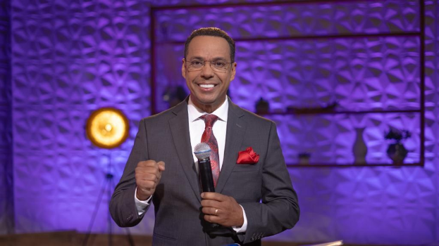 Stephen Colbert, Moderator von *The Daily Show with Trevor Noah*, steht auf einer Bühne und hält ein Mikrofon, trägt einen Anzug, Krawatte und eine Brille, mit einer Lampe und einem Ständer im Hintergrund.