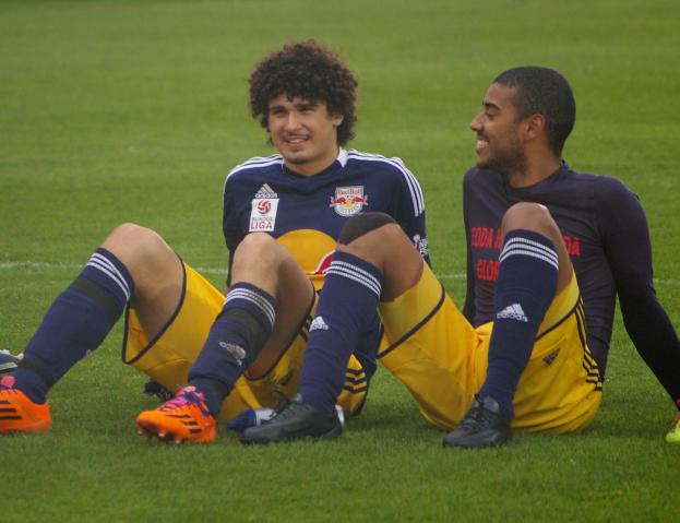 Zwei Fussballspieler sitzen auf dem Boden mit gekreuzten Beinen, tragen Sportbekleidung und lächeln sich an.