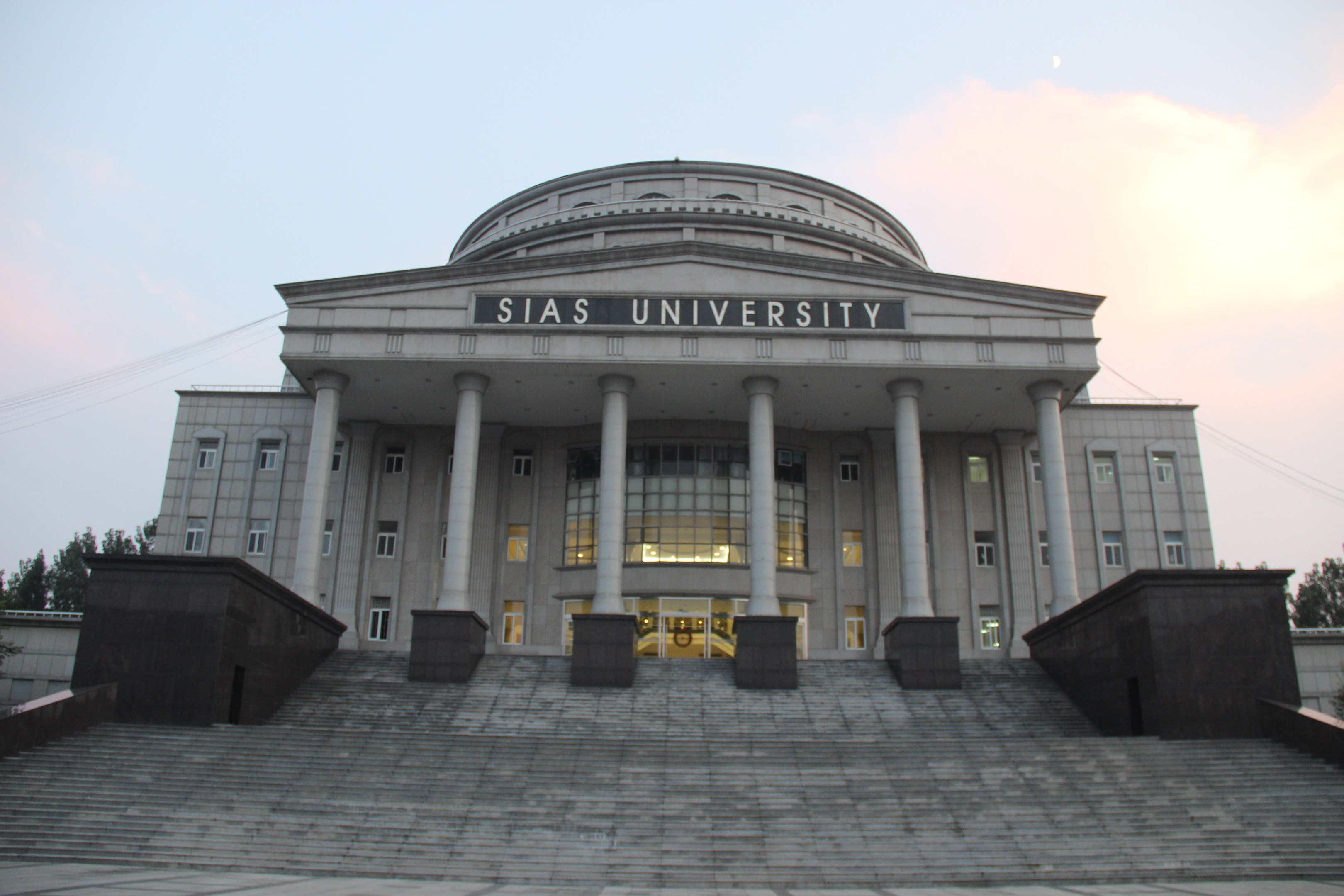 Large building with pillars, windows, doors, and steps, surrounded by trees under a clear blue sky, with the university's name visible on the facade.