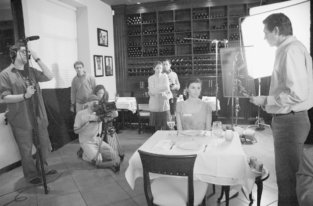 Ein Filmset mit einem Mann rechts, einer Frau an einem Tisch mit Tellern, Gabeln und Gläsern in der Mitte und zwei Personen links, die eine Kamera bedienen, sowie Regalen, Rahmen und einem Bildschirm im Hintergrund.