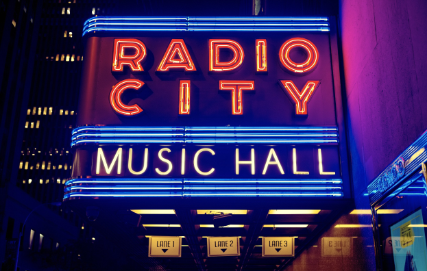 Hell erleuchtetes Radio City Music Hall in New York City mit hohen Gebäuden im Hintergrund und einem "Radio City"-Schild auf der rechten Seite mit zusätzlichen Tafeln darunter.