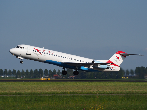 Austrian Airlines Embraer ERJ-190 aircraft taking off from Frankfurt Airport, with green grass, trees, buildings, and sky visible in the background.