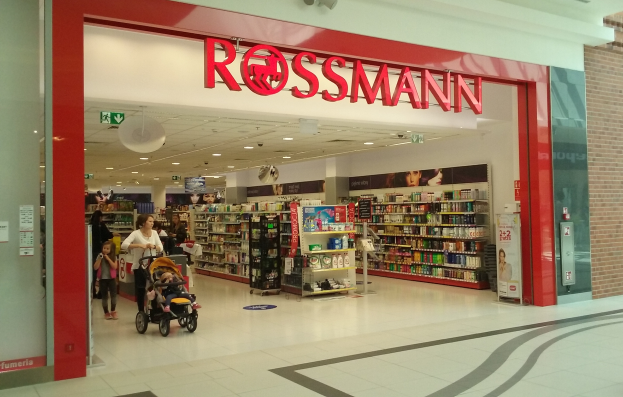 Ein Rossmann-Laden in einem Einkaufszentrum mit Menschen auf dem Boden, einer hält einen Kinderwagen, und Regale voller verschiedener Gegenstände, beleuchtet von Deckenlampen und Schildern mit Text.
