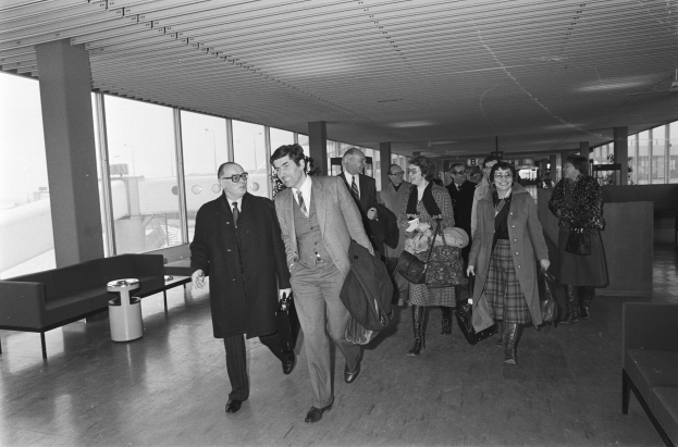 Schwarz-weiß-Foto von Menschen, die durch einen Flughafen gehen und Koffer tragen, mit Sofas, einem Müllcontainer, Säulen und Glasfenstern im Hintergrund.