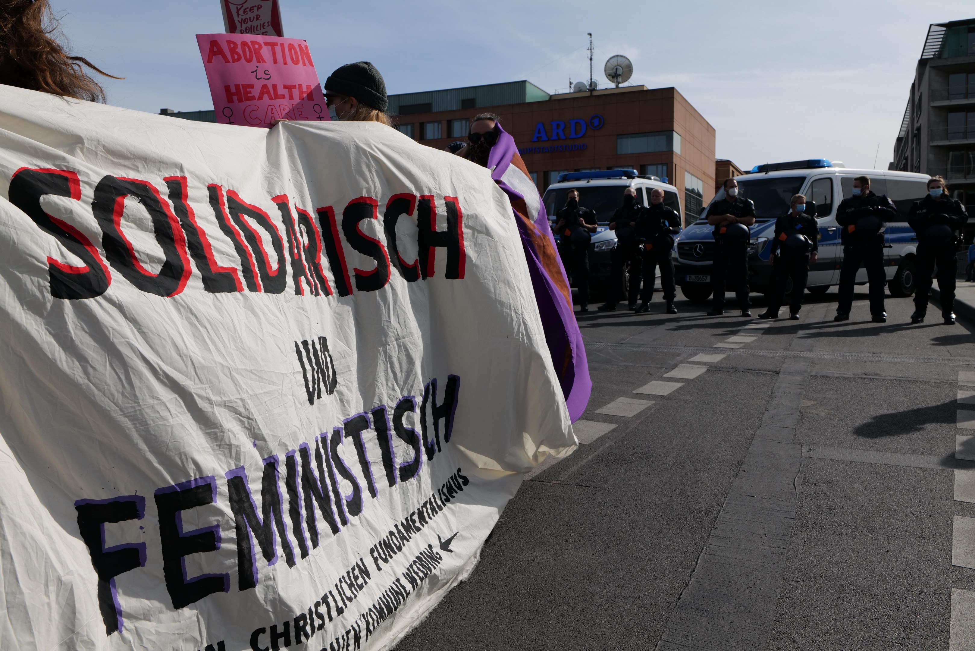 Gruppe von Menschen marschiert auf einer Straße und hält eine 'Solidarität und Feminismus'-Schleife, mit parkenden Fahrzeugen, Gebäuden, einer Schüsselantenne und einem klaren blauen Himmel im Hintergrund.