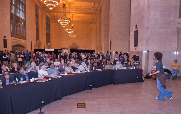 Eine Konzertszene mit einer Menge, einem Mann, der auf der rechten Seite Gitarre spielt und singt, einer Wand und Fenstern im Hintergrund, Lichtern oben und einem Boden unten.