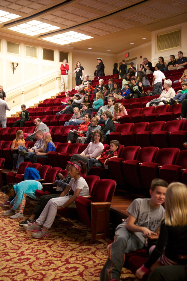 Eine große Gruppe von Menschen, darunter Kinder, die auf Stühlen und Stehplätzen in einem Theater sitzen und eine beleuchtete Wand und Decke im Hintergrund.