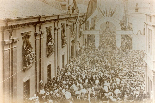 Black and white photo of a large crowd celebrating a victory parade, walking down a street lined with buildings, passing under an arch in the background.