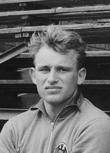 Black and white photo of a man in a jacket standing in front of a pile of wooden planks, believed to be a legendary German footballer.