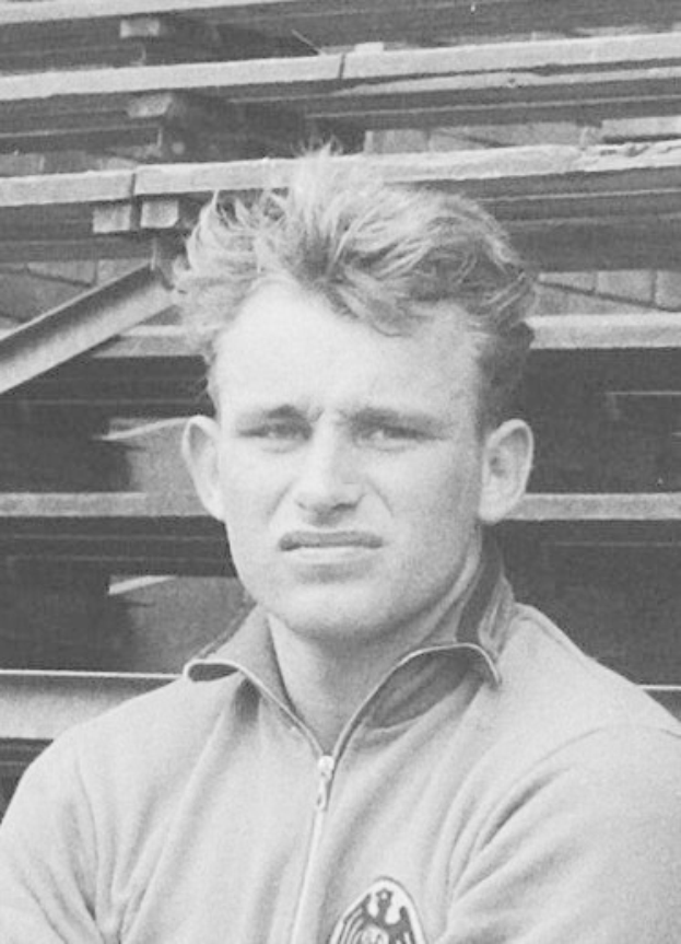 Black and white photo of a man in a jacket standing in front of a pile of wooden planks, believed to be a legendary German footballer.