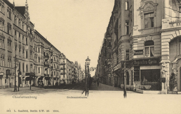 Schwarz-weißes Foto einer Straßenansicht in Charlottenburg, Deutschland, mit Gebäuden, Straßenlaternen, Schildern, Motorfahrzeugen, Fußgängern und Gittern unter einem klaren Himmel.