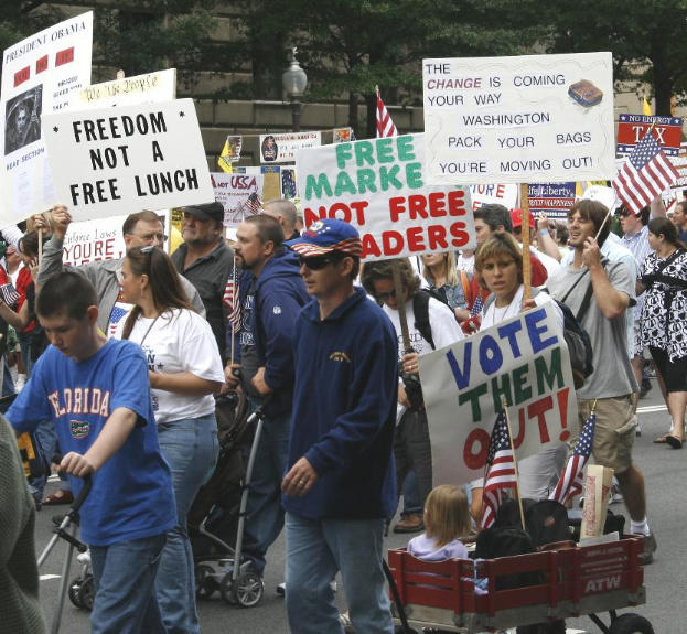 Eine Gruppe von Menschen, die auf einer Straße mit Schildern geht, mit Kindern in einem Wagen vorne und einem Baum, einem Laternenpfahl und einem Gebäude im Hintergrund.