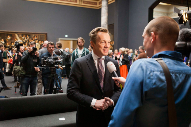 Ein Mann im Anzug und mit Krawatte spricht mit einem Reporter und hält ein Mikrofon in der Hand, im Hintergrund ist eine Menschenmenge zu sehen, einige halten Kameras, im Vordergrund ein Sofa und eine Wand mit einem Foto Rahmen und Lampen dahinter.
