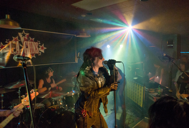 Eine Frau in einer schwarzen Jacke singt auf der Bühne in ein Mikrofon, während ein Mann mit Sonnenbrille und Stirnbinde ein Instrument spielt und ein Plakat mit 'LA Guns' im Hintergrund zu sehen ist, unter Diskoleuchten und Hängelampen.