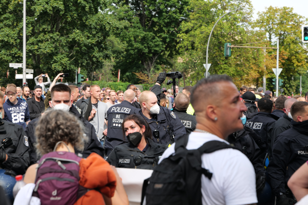 Eine große Gruppe von Menschen, einige mit Masken und Rucksäcken oder Kameras, steht vor einer Gruppe von Polizisten, mit Laternenmasten, Verkehrsampeln, Schildern, Bäumen und einem klaren blauen Himmel im Hintergrund.