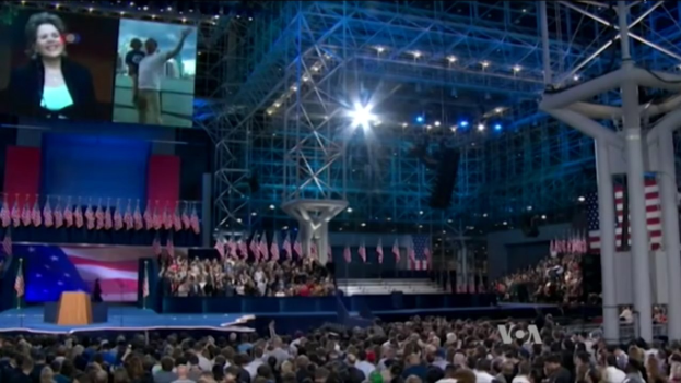 Eine große Zuschauermenge sitzt einer Bühne mit einem Podium, einem Mikrofon, Flaggen, einem Overhead-Bildschirm und einer Lichtanlage gegenüber, während es scheint, als fände ein Präsidentschaftsdebatt statt.