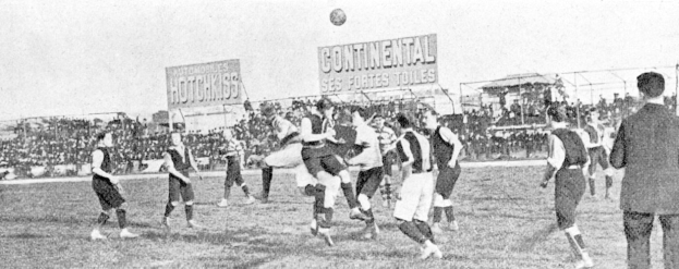 Ein Schwarz-Weiß-Bild einer Gruppe von Männern, die auf einem Feld Fußball spielen, mit einem Ball in der Luft und Zuschauern im Hintergrund, mit Texttafeln, die wahrscheinlich Matchdetails angeben.