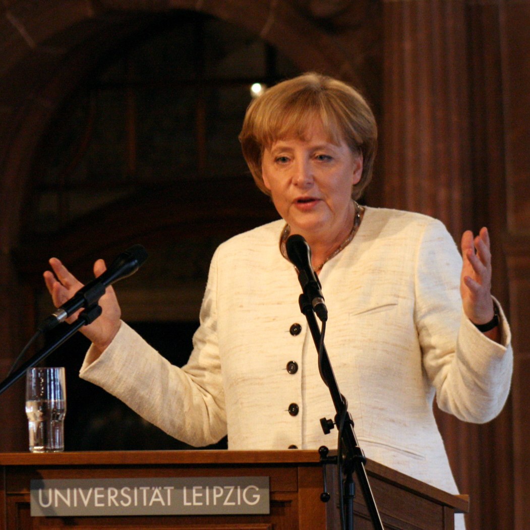 German Chancellor Angela Merkel delivering a speech at the University of Leipzig, standing at a podium with a microphone and a glass of water.