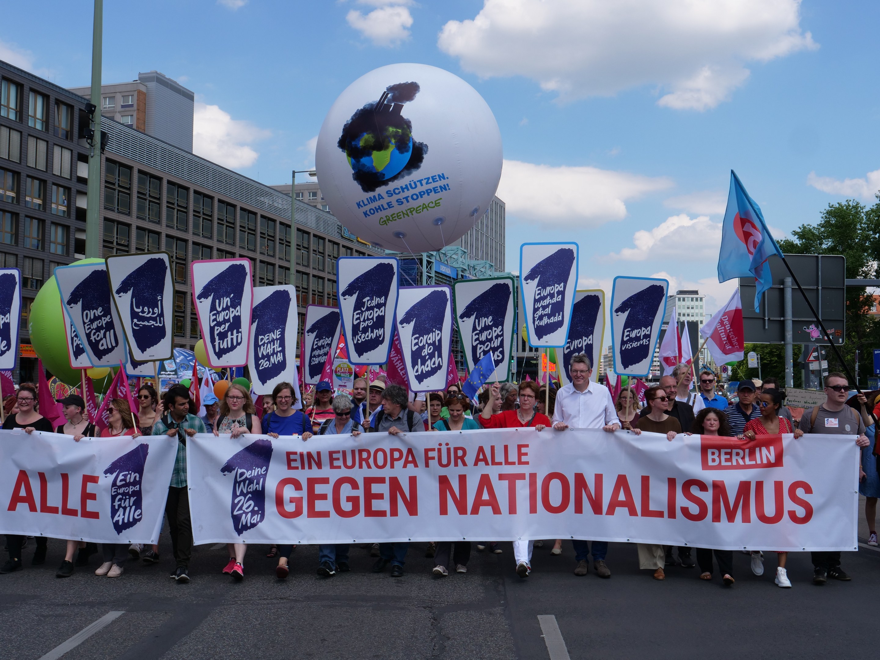 A group of people marching down a street, holding a banner reading "Ein Europa für alle Gegen Nationalismus" and flags, with buildings, trees, and a cloudy sky in the background.