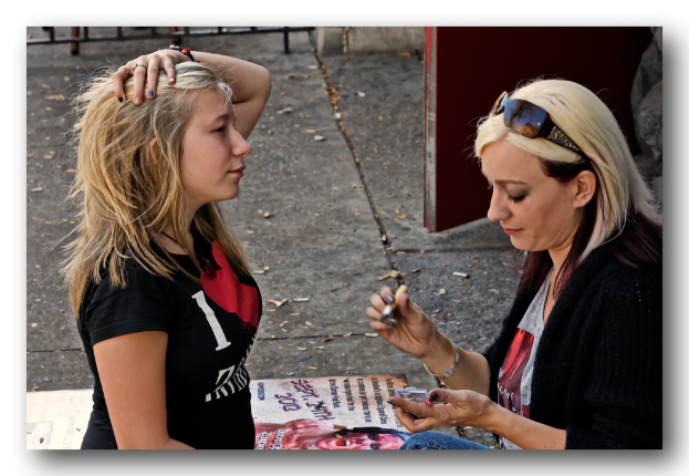 Eine Frau in einem schwarzen T-Shirt sitzt links, während eine andere Frau rechts ein Objekt hält, mit einer Tür, einem Geländer und einer Wand im Hintergrund sowie Zigaretten, einem Banner mit Text und einem Banner auf dem Boden.