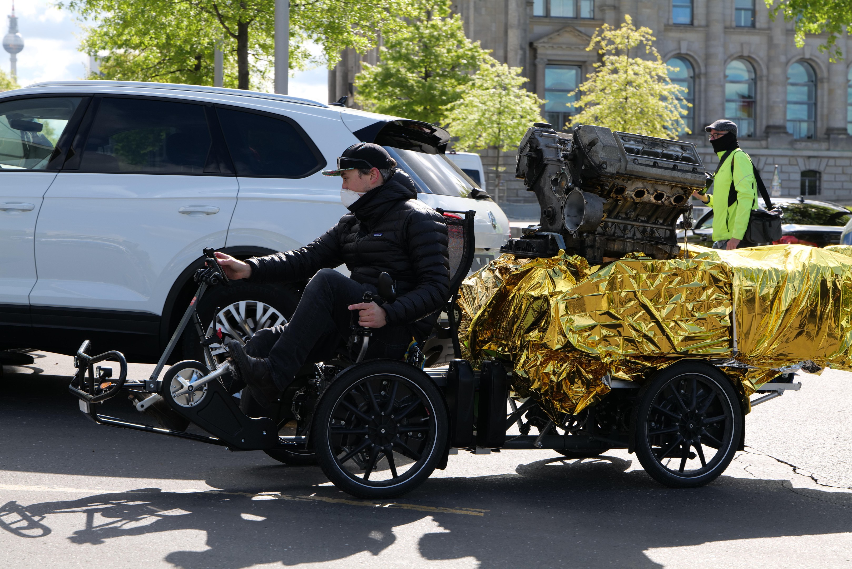 A man in a wheelchair with a large engine on the back, holding an object, on a road surrounded by vehicles, trees, buildings, poles, and a clear blue sky.