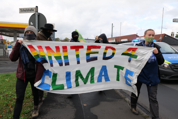Eine Gruppe maskierter Personen mit einer 'United Climate'-Schleife vor einer Tankstelle, mit Fahrzeugen auf der Straße, Gras, Schildern, Gebäuden, Bäumen und einem bewölkten Himmel im Hintergrund.