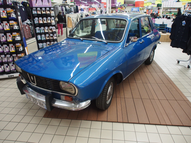 Blauer Wagen in einem Bekleidungsgeschäft mit Kleiderstangen, Menschen, Kartons, Schildern und Deckenleuchten.