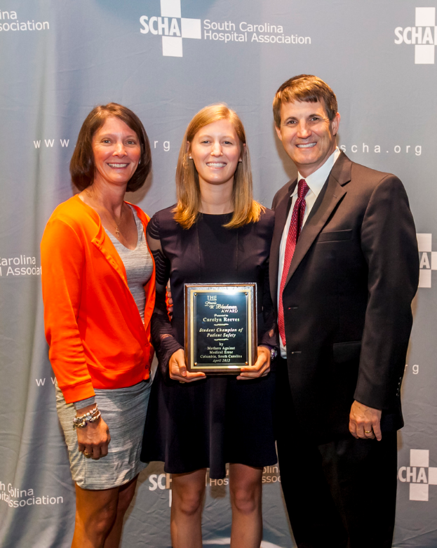 Drei Personen, zwei Frauen und ein Mann, die gemeinsam mit einer Frau, die eine Auszeichnung hält, vor einem grauen Hintergrund stehen.