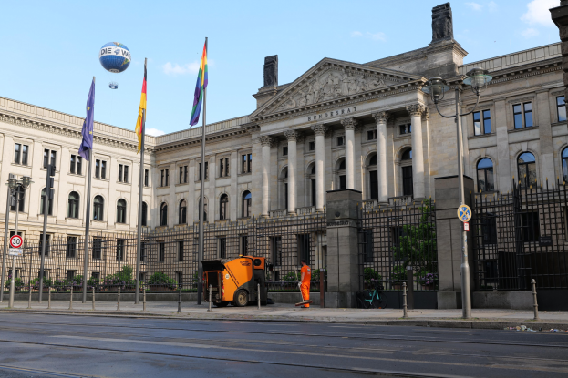Large modern building with many windows, identified as the Bundestag in Berlin, Germany, surrounded by street infrastructure, pedestrians, flags, and trees under a cloudy sky.