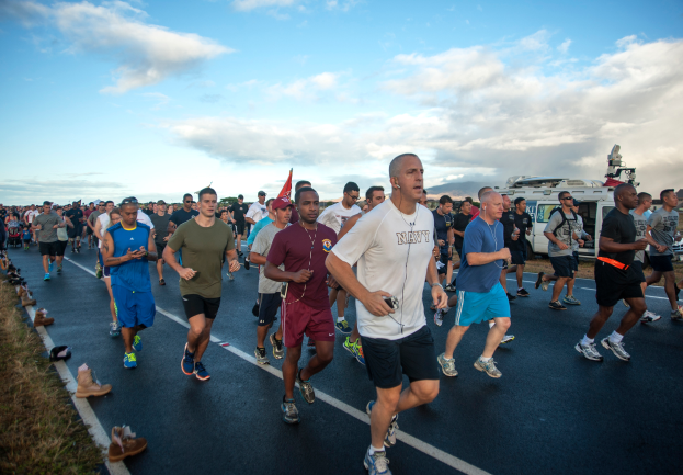 Eine Gruppe von Menschen, die bei einem Marathon auf einer Straße laufen, mit Gras und Schuhwerk im Vordergrund und Fahrzeugen, Bäumen, Hügeln und einem bewölkten Himmel im Hintergrund.