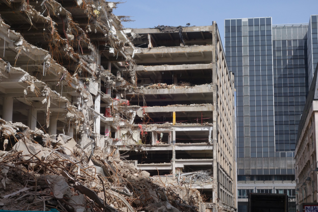 A large building demolished in a city, surrounded by other structures, street elements, vehicles, trees, and a clear sky.
