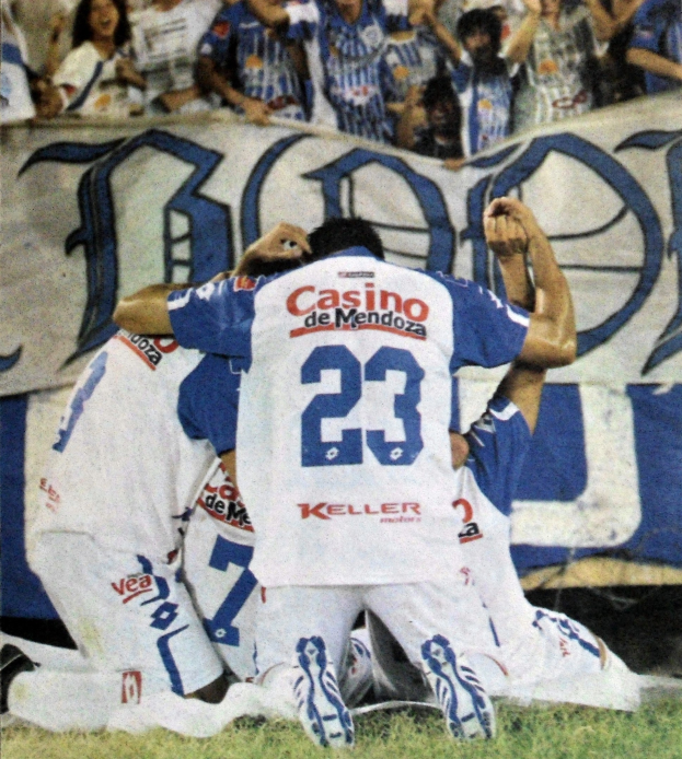 Gruppe von Fußballspielern, die ein Tor feiern, auf dem Boden mit einer jubelnden Menge im Hintergrund und einem Banner mit Text dahinter.