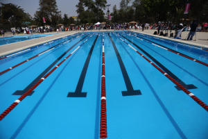 Ein großes Schwimmbad mit Bahnentrennungen, umgeben von Menschen mit Taschen und anderen Gegenständen, sowie Zelten, Bannern, Stangen, einer Straßenlaterne und einem bewölkten Himmel im Hintergrund.