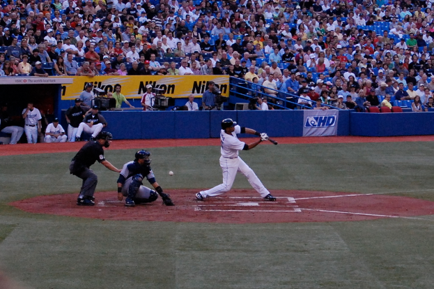 Ein Mann hält einen Baseballschläger mit einem Ball hinter sich, während mehrere Personen auf Stühlen sitzen und andere Kameras halten, mit Werbetafeln und Metallstäben davor.