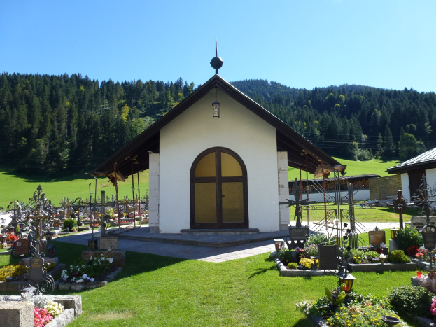 Small chapel with a door and windows, surrounded by a grassy field, cemetery, plants with flowers, stones, and poles, set against trees and a clear blue sky.