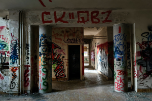 Abandoned building with graffiti-covered walls, pillars, exposed pipes, and a visible floor, featuring a door in the background.