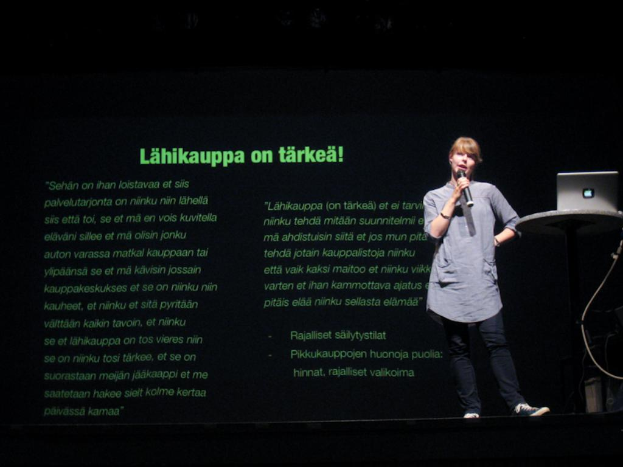 Eine Frau steht auf einer Bühne und spricht, mit einem Apple-Laptop auf einem Tisch neben ihr und einem Projektor, der Inhalt hinter ihr anzeigt.