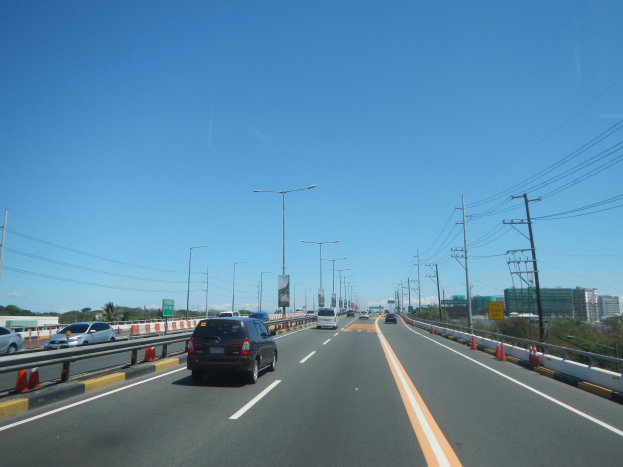 Eine Autobahn mit Autos, Verkehrskegeln, Laternenmästen, Strommästen mit Drähten, Texttafeln, Bäumen, Gebäuden und einem klaren blauen Himmel.