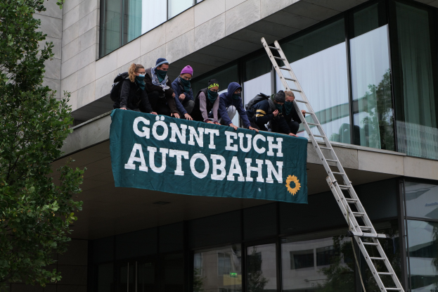 Gruppe von Menschen auf einem Gebäude mit einem Banner, mit einem Baum links und Glasfenstern im Hintergrund, wahrscheinlich protestierend für die deutsche Regierung.