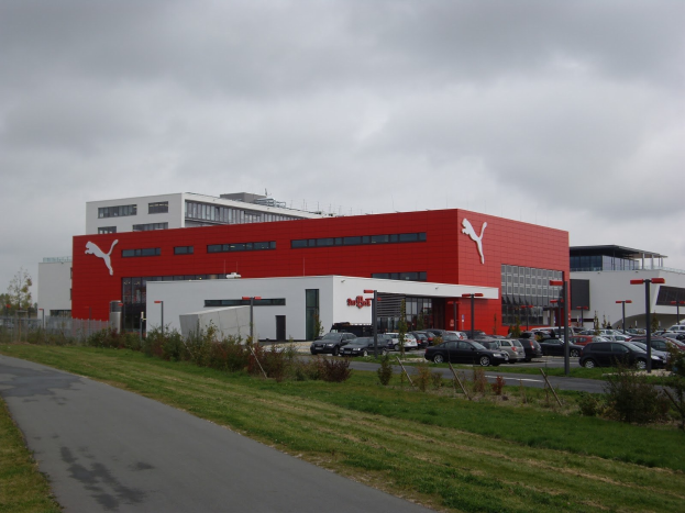 Fabrikgebäude von Puma in Deutschland mit Fahrzeugen auf der Straße, Straßeninfrastruktur, Vegetation und einem bewölkten Himmel.