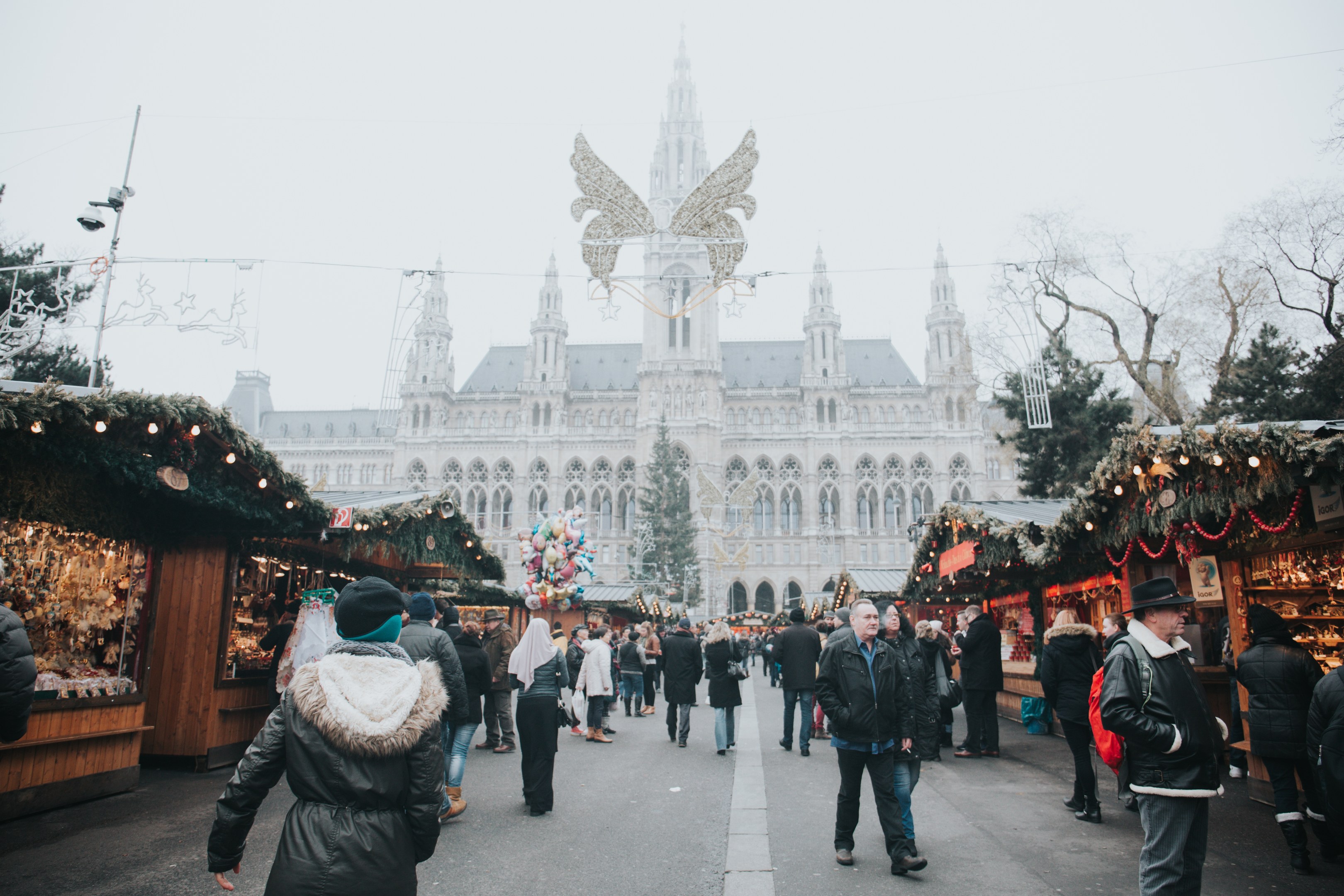 Ein belebter Weihnachtsmarkt in Wien, Österreich, mit Menschen, die herumlaufen, Ständen, die mit Lichtern und festlichen Artikeln geschmückt sind, einem Gebäude mit Fenstern im Hintergrund, Bäumen und einem klaren blauen Himmel.
