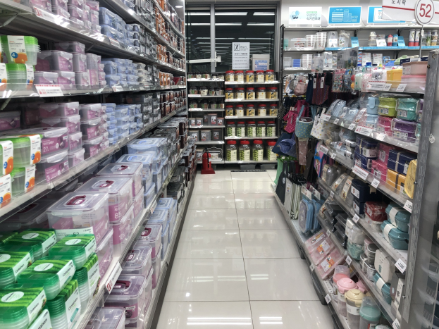 Ein unordentlicher chinesischer Supermarktgang mit Regalen voller Kartons, Flaschen, Taschen und anderen Produkten, beleuchtet von Deckenlampen und gekennzeichnet durch Texttafeln und einen sichtbaren Feuerlöscher.