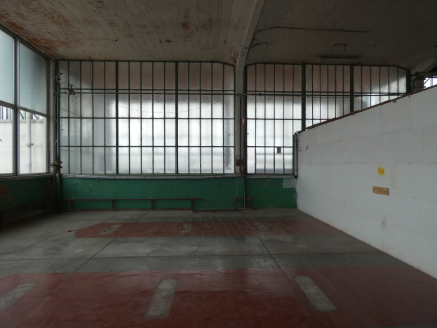 Leerer Raum mit rotem Boden, weißer Wand auf der rechten Seite, Metallstangen und Fenstern, die an eine Gefängniszelle erinnern.