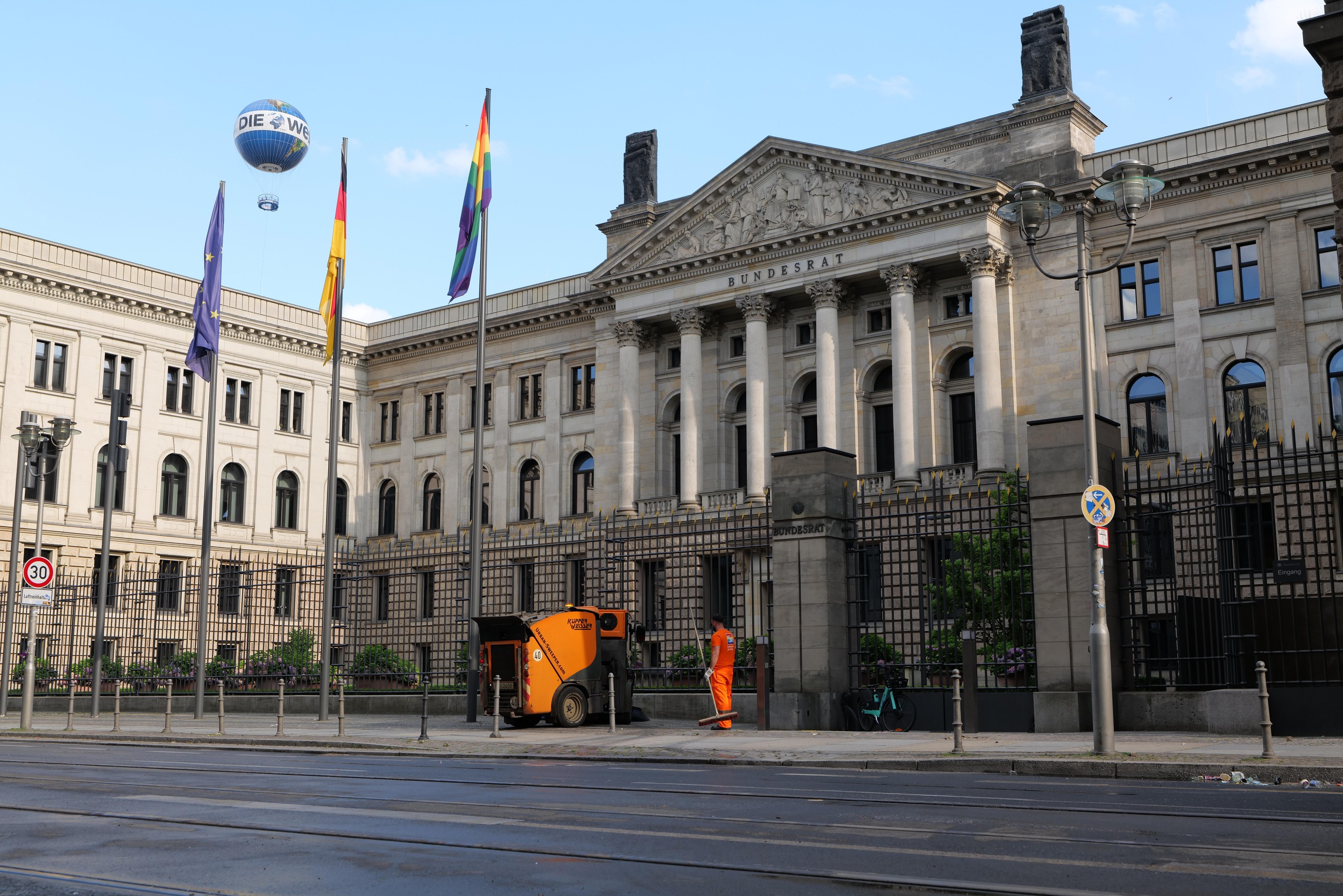 Large modern building with numerous windows, identified as the Bundestag in Berlin, Germany, surrounded by street infrastructure, vehicles, pedestrians, and trees under a cloudy sky.
