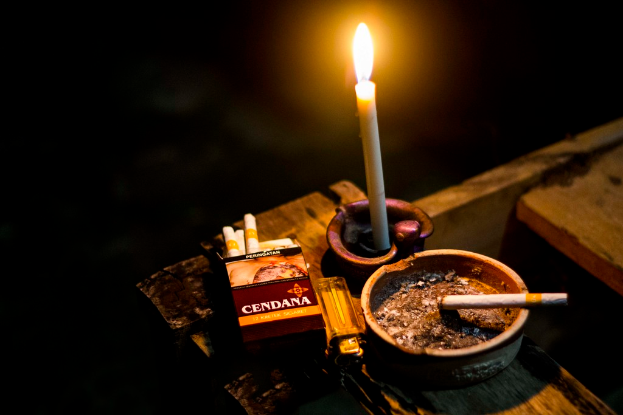 Eine Kerze, Zigaretten, eine Schachtel, ein Feuerzeug und ein Behälter sind auf einer Oberfläche angeordnet.