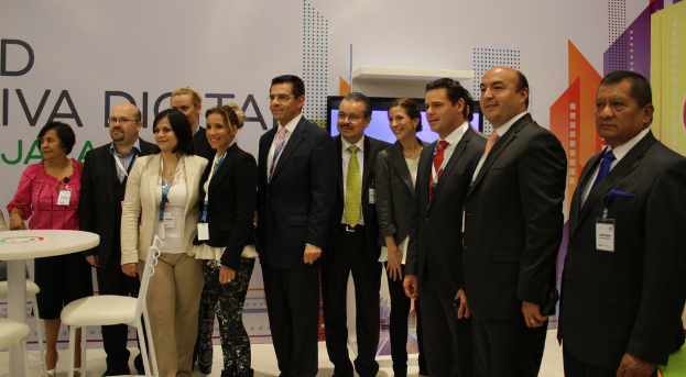 Eine Gruppe von Menschen in Anzügen, die in der Mitte stehen, mit einer Gruppe von lachenden Frauen in der Nähe, einem Tisch, Stühlen und anderen Gegenständen im Vordergrund und einem Banner mit Text und einem digitalen Bildschirm im Hintergrund.