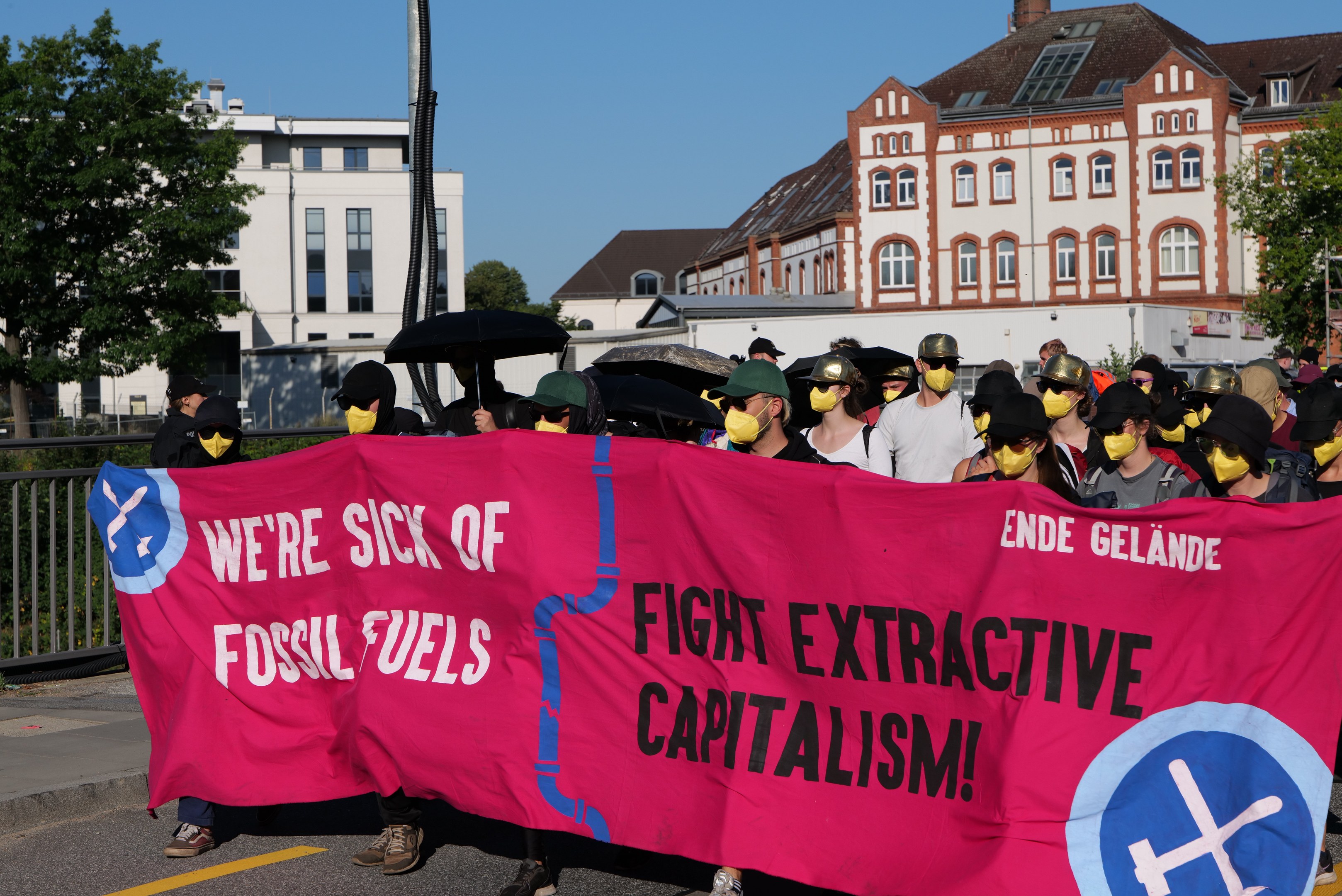Eine Gruppe von Menschen in Masken mit einem pinken Banner mit der Aufschrift "Wir haben genug von fossilen Brennstoffen, bekämpft den extraktiven Kapitalismus" auf der Straße, einige mit Schirmen, vor einem Hintergrund von Gebäuden, Bäumen, einem Pfahl und einem klaren blauen Himmel.