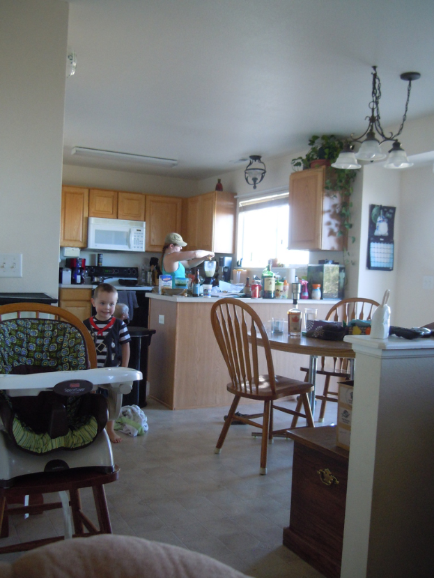 Eine Frau arbeitet in der Küche, während ein kleines Kind in der Nähe spielt; ein Tisch mit Gläsern, einer Flasche und Essen befindet sich rechts daneben, umgeben von Stühlen. Die Küche enthält Gläser, Geschirr, einen Mixer, Flaschen, Schränke, einen Kronleuchter, eine Pflanze und eine Deckenlampe.