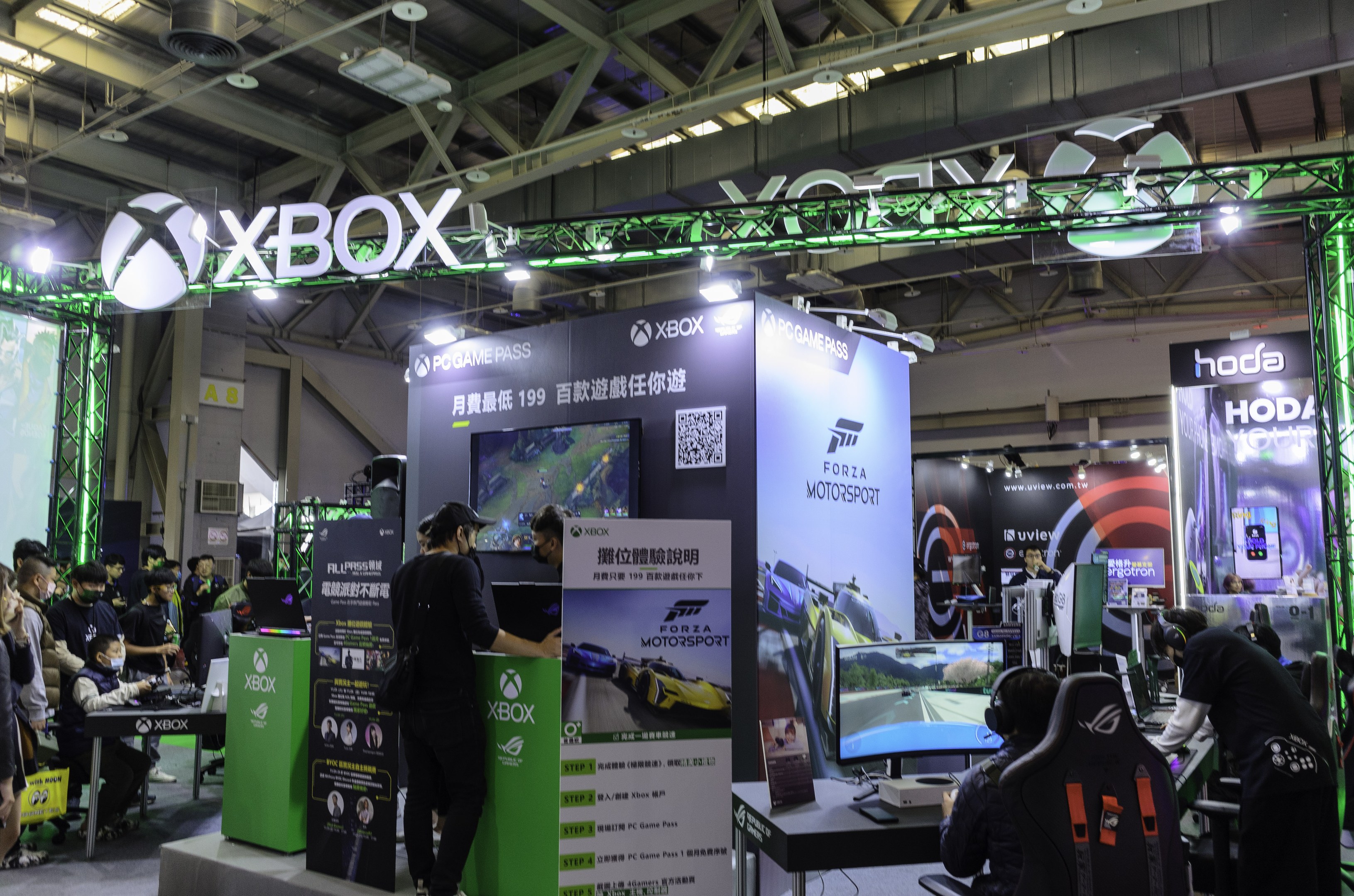 Eine Gruppe von Menschen an einem Messestand mit dem Motto 'Forza Motorsport 5 China', mit Tischen, Monitoren, Tastaturen, Bannern und einem Metalldach im Hintergrund.